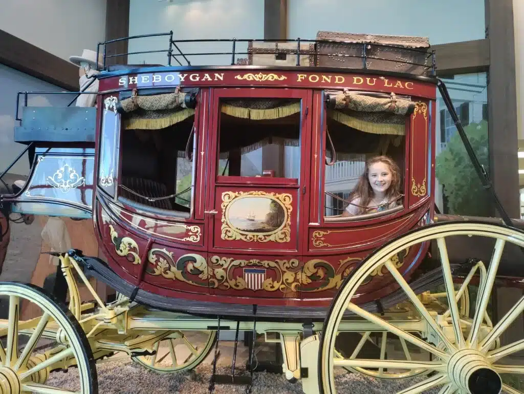 A girl in pink shirt riding a horse-drawn red carriage at Wade House Greenbush Wisconsin