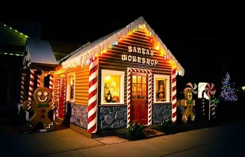 House with snow-covered rooftop with an inscription that says Santa's Workshop, red front door and a red and white striped siding.