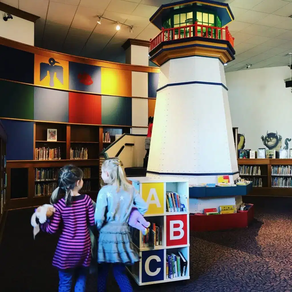 A close shot of the wooden lighthouse with stacks of books in the bottom and two kids walking