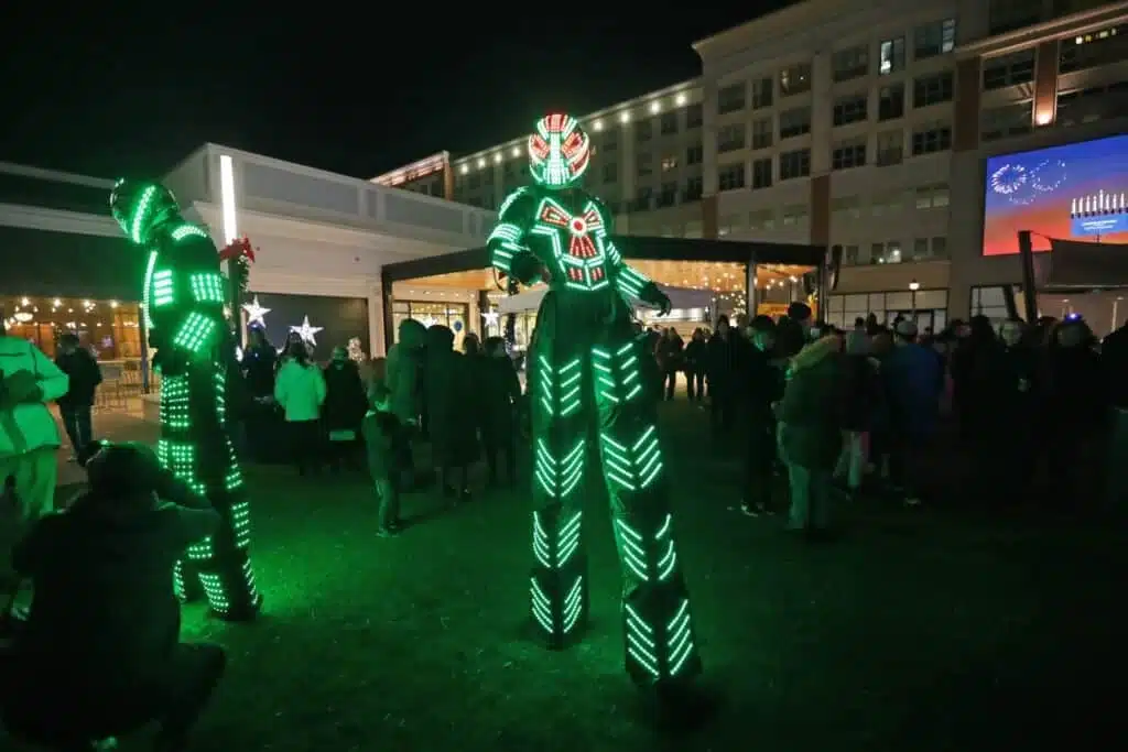 A large celebration in front of a white building with lots of people celebrating and two neon clad robots.