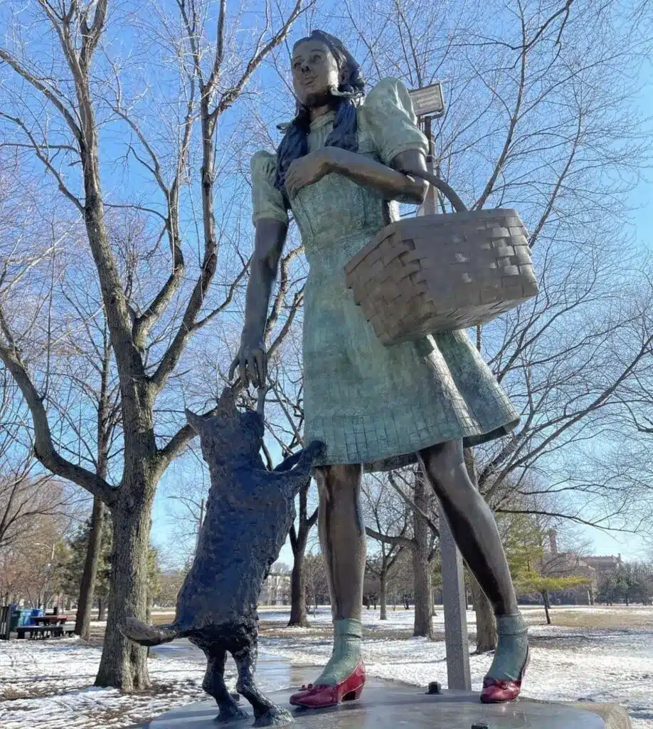 A tall bronze statue of Dorothy and Toto from “The Wizard of Oz” standing in Oz Park in Chicago, with winter trees and light snow on the ground.