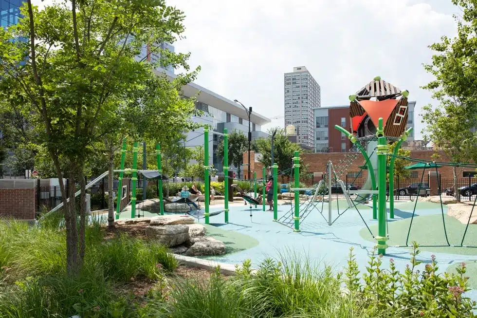 A modern Chicago playground at Prairie District Park featuring bright green climbing structures, soft-surface flooring, and lush landscaping with city buildings in the background.