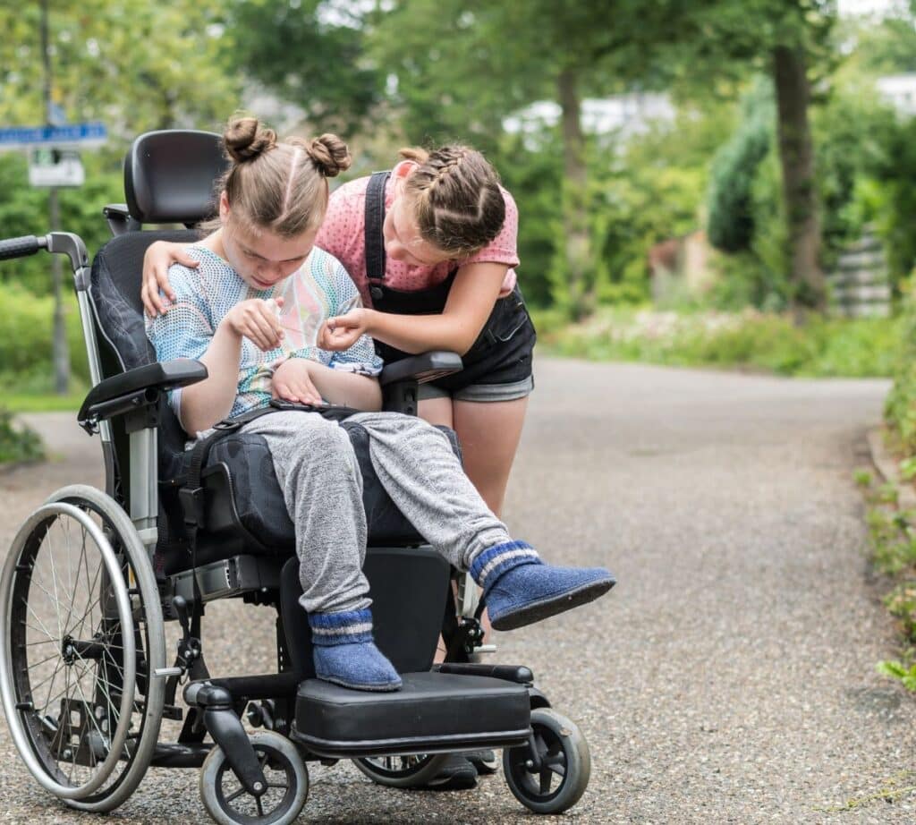A child gently interacting with another child who is seated in a wheelchair along a quiet outdoor path, offering support and connection.