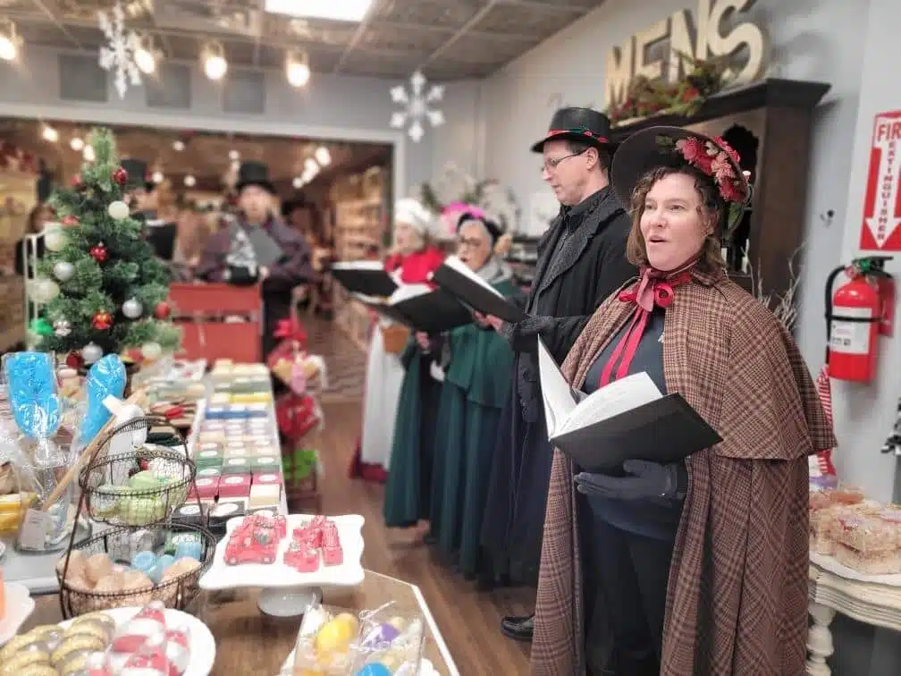 Carolers dressed in 19th century period costumes holding a songbook and singing in front of a yuletide table spread