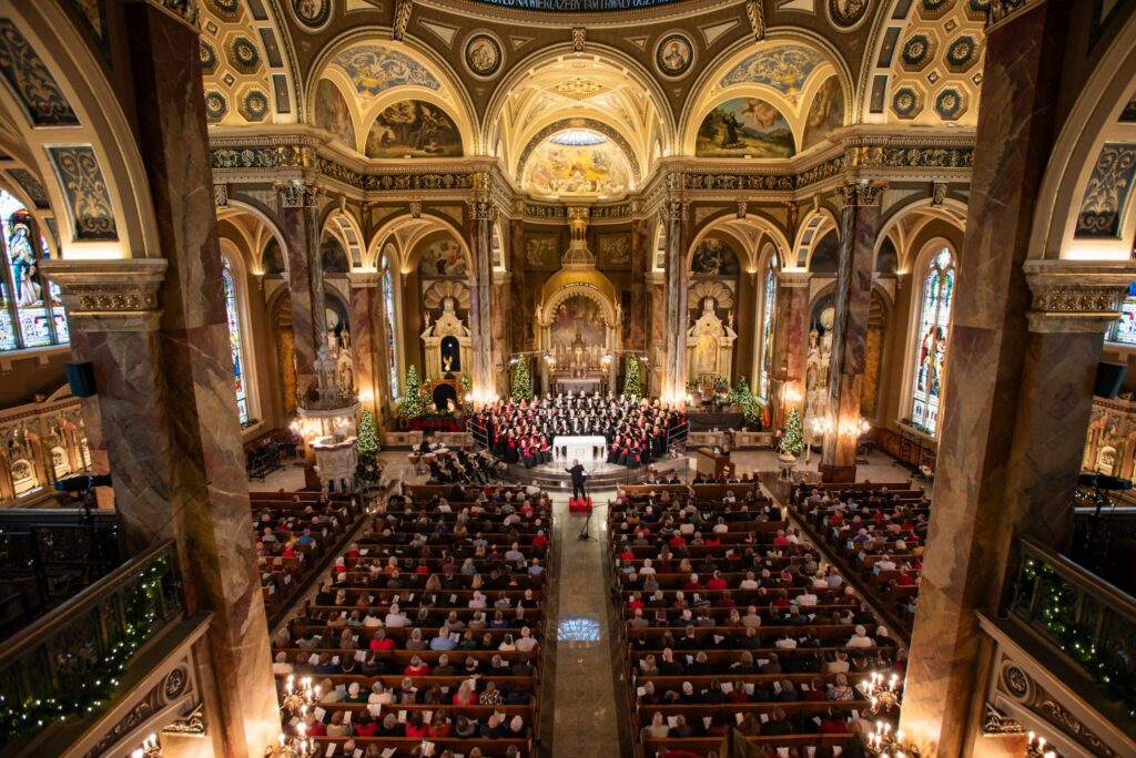 A full choir performs inside the beautifully ornate Basilica of St. Josaphat in Milwaukee, surrounded by Christmas trees, golden arches, and stained-glass windows as an audience fills the pews below.