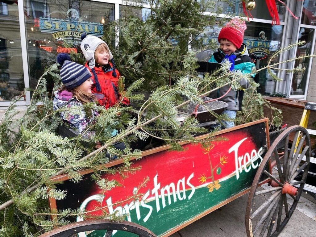 Three smiling kids on top of a cart with label Christmas Trees painted on its side at the Charlie Brown Christmas tree lot in Milwaukee Public Market,