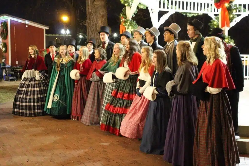 Carolers in 19th century historical costumes that includes capes and white Victorian muffs for women and tall hats and scarves for men.