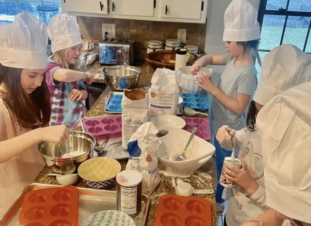 Young children wearing chef hats mixing ingredients and preparing donut batter at a kitchen counter.
