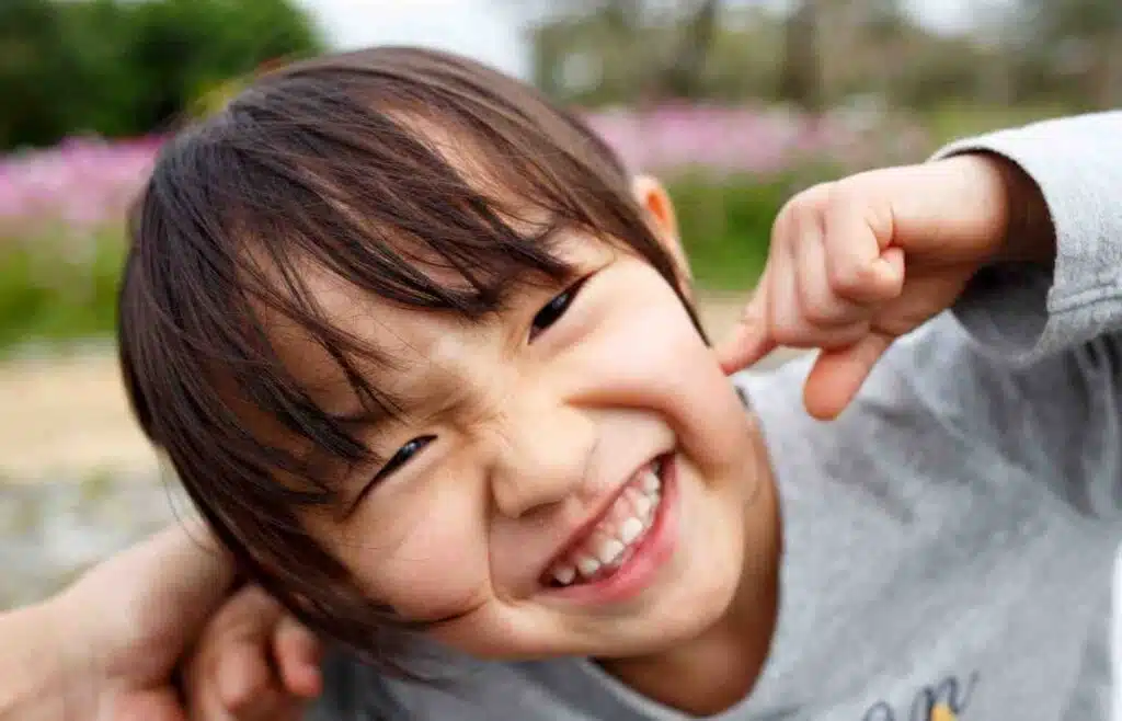 A young child with a big smile, playfully pulling at their cheeks with their fingers, expressing pure joy and laughter.