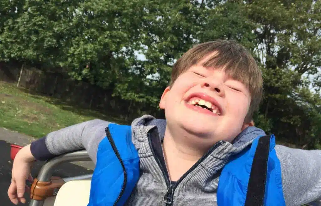 A child laughing joyfully outdoors, sitting with arms wide open and eyes closed on a sunny day.