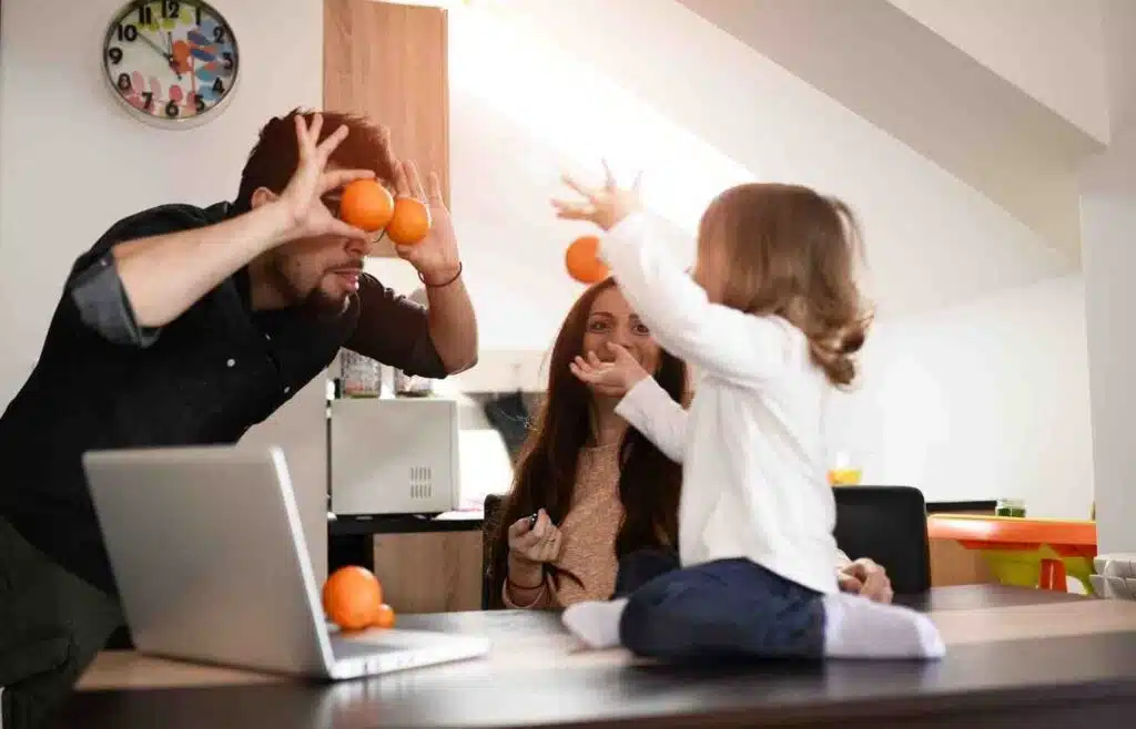 Family laughing in kitchen while dad makes orange glasses and child throws fruit