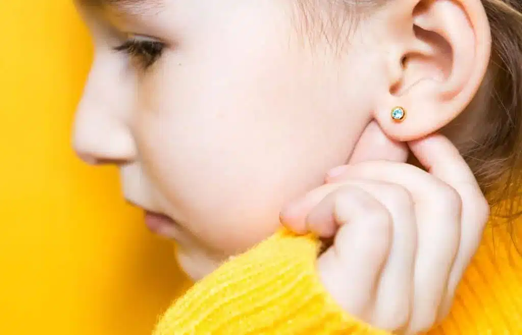 Child with newly pierced ear touching small stud earring against bright yellow background.