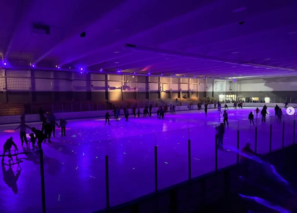 Skaters of all ages glide under purple glow lights during a Glow Skate event at Eble Ice Arena, with colorful dots reflecting across the ice.