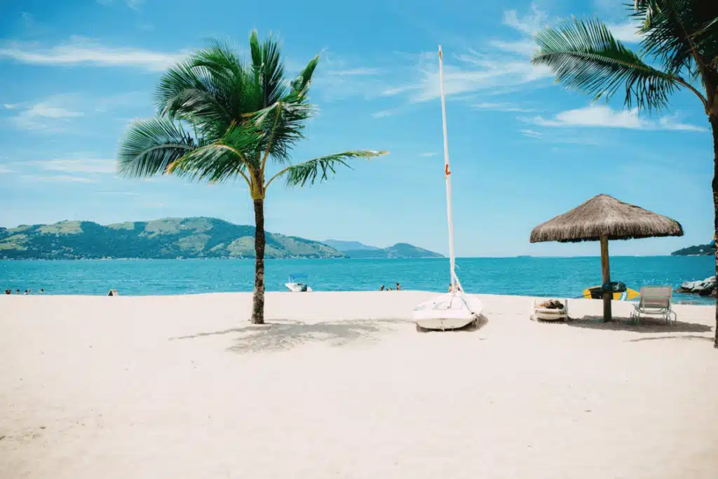 A beach cabana with lounge chairs beside a small sailboat and coconut trees