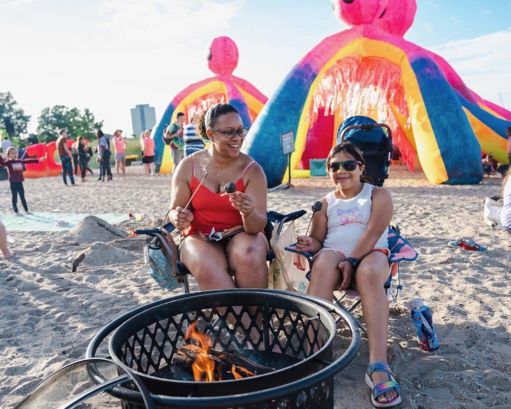 Mom and daughter roast marshmallows over a fire pit on the beach at South Shore Park in Milwaukee with giant inflatable octopus behind them. 