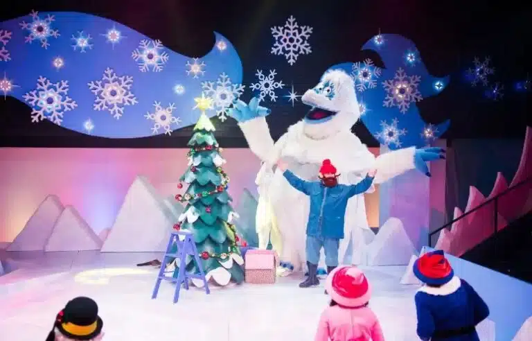 A stage with an actor performing and behind him is a giant white yeti and a Christmas tree