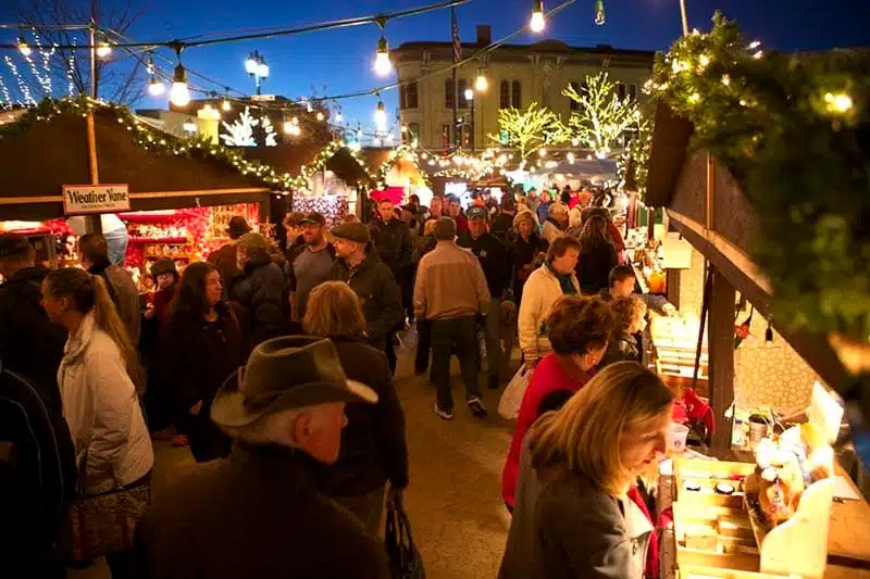 People shopping for holiday gifts at the Oconomowoc German Christmas Market with stalls decorated with lights and tree branches at dusk