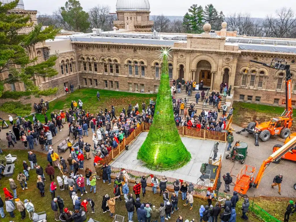 People gathering around the green glass Christmas tree in front of a building and some construction cranes placed on the right side.