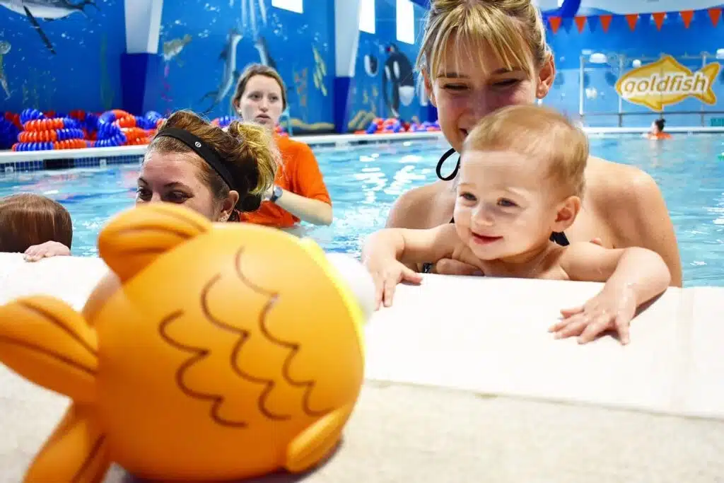 A toddler learning how to swim together with the coach with logo of Goldfish Swim School in the background