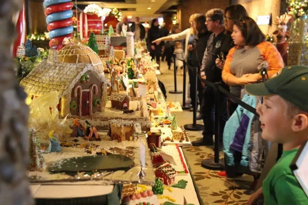 A boy with a green shirt and a cap is happily looking at a tall gingerbread house and one person is pointing at a gingerbread house.
