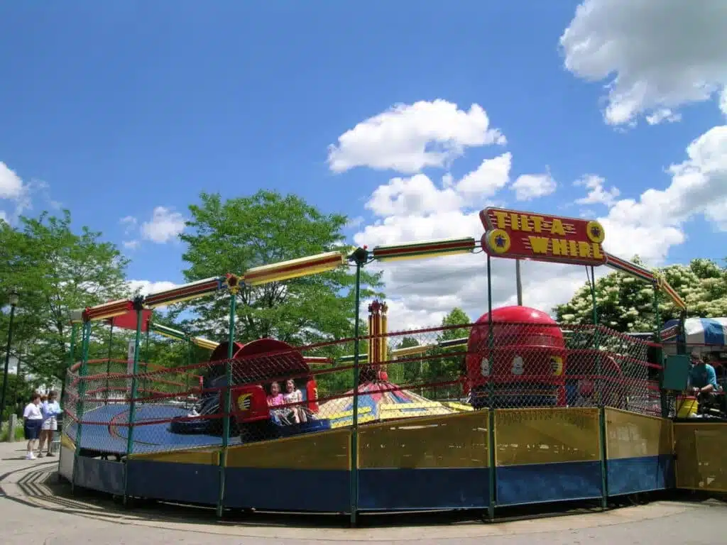 Children ride the classic red and yellow Tilt-A-Whirl on a sunny day, surrounded by trees and bright blue sky.