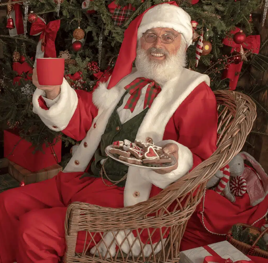 Santa sitting on a wicker chair holding a red ceramic mug and a plate of Christmas cookies.