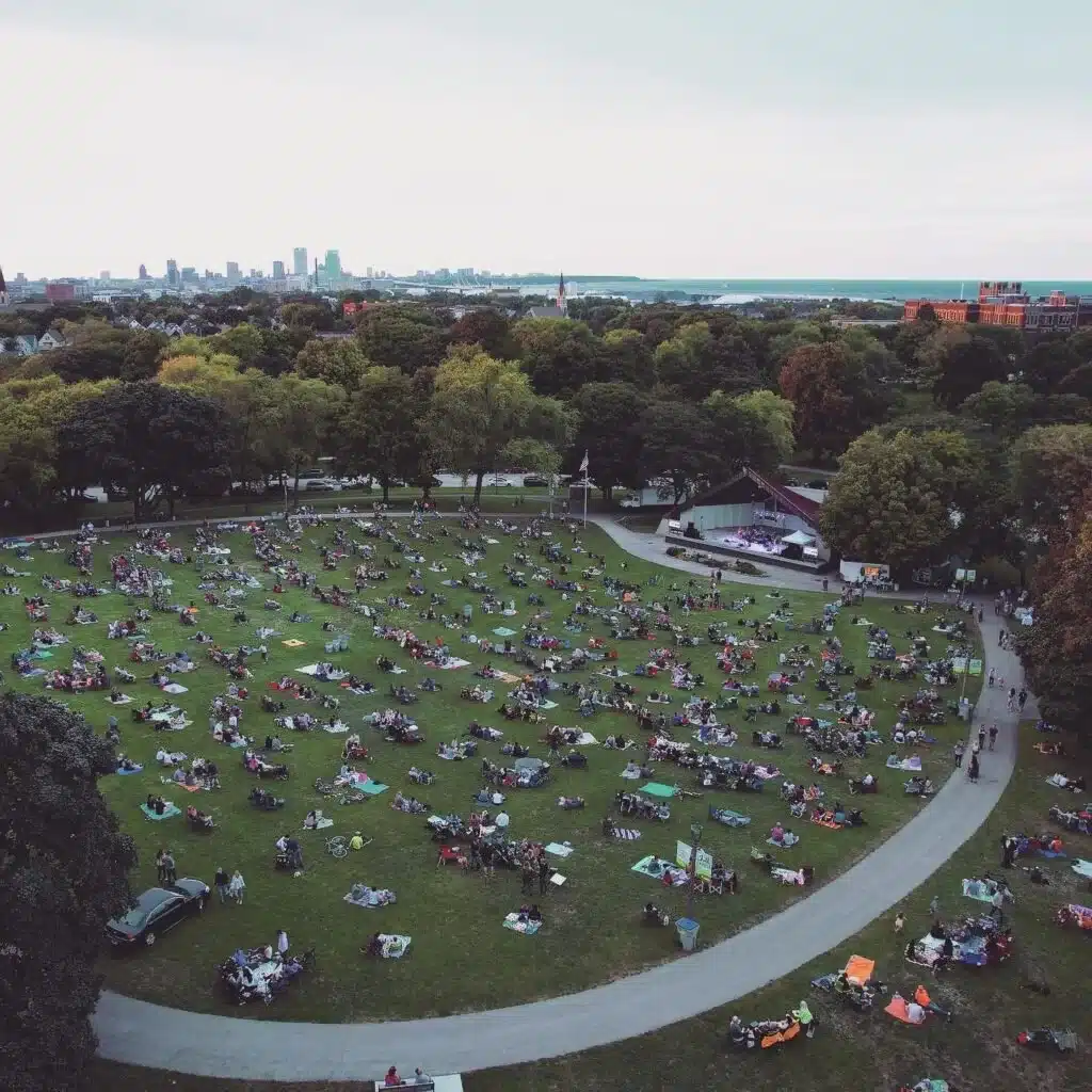 A wide aerial view of Chill on the Hill at Humboldt Park in Milwaukee, with dozens of families relaxing on blankets across a large circular lawn.