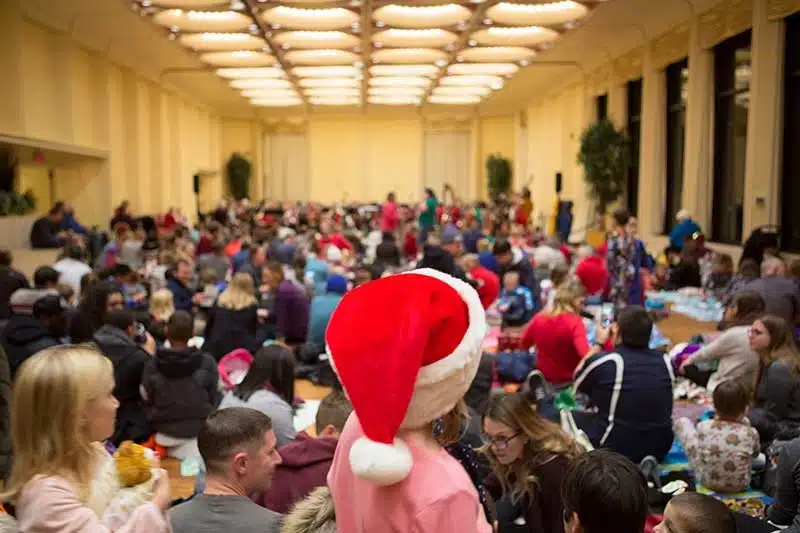 Families and kids sitting inside a large function hall and child in pink shirt with santa hat,