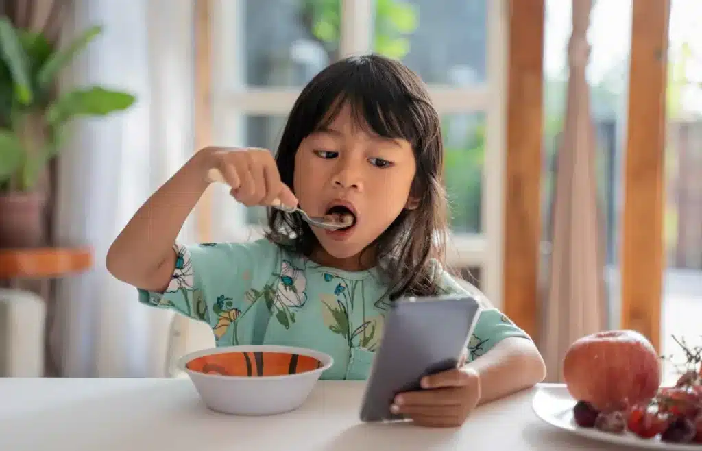 Young child eating breakfast while looking at a smartphone.