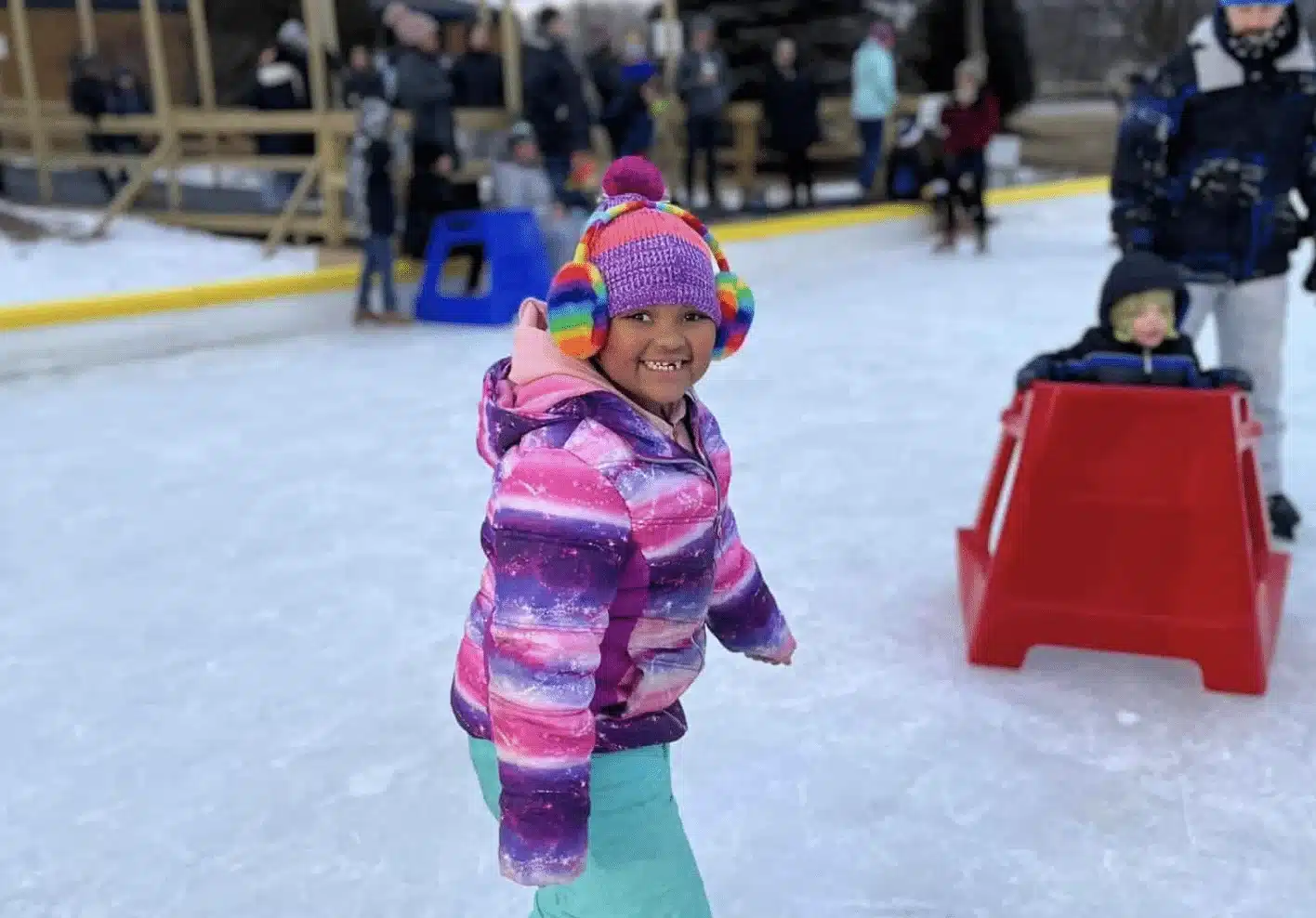 Child in purple jacket and rainbow muffe ice skating with skate aids at Konkel Park
