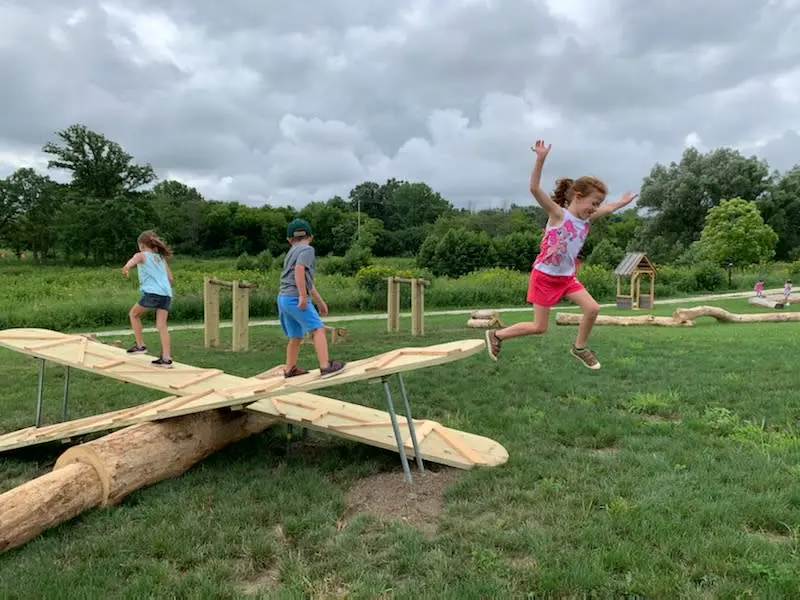 Three kids are playing on a giant see-saw with some hurdles and a small playhouse at the back