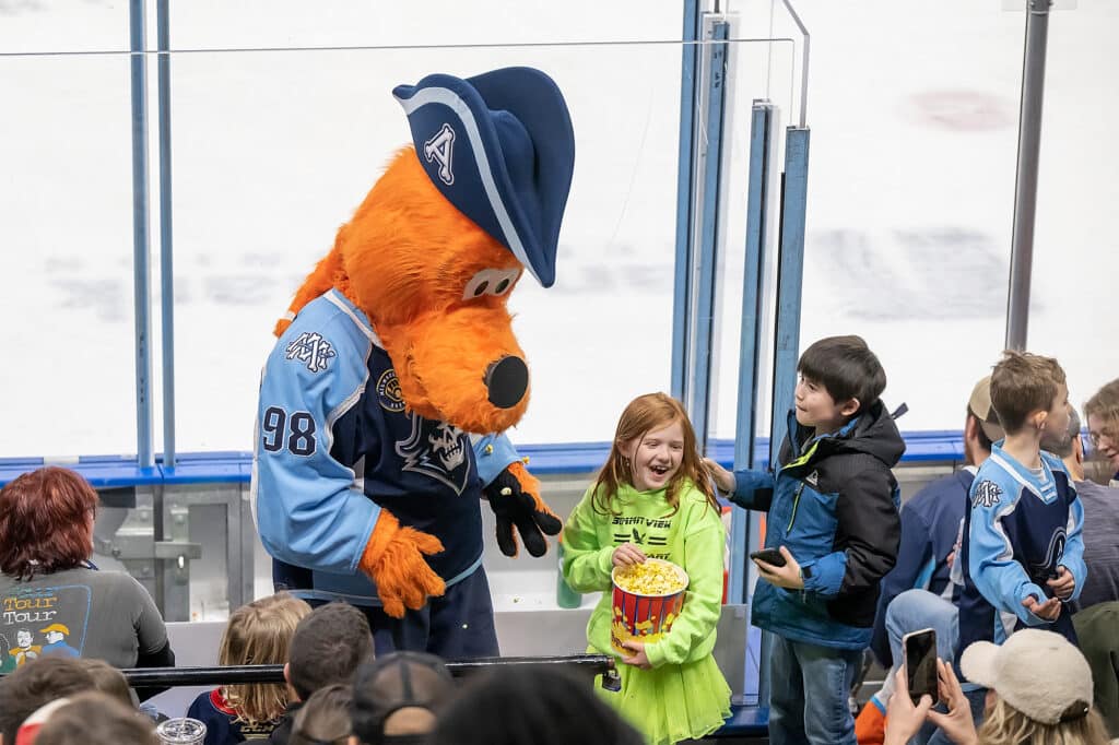 A young girl laughing while holdling a tub of popcorn with a young boy while the Milwaukee Admirals mascot is looking on