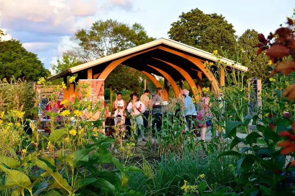 Families gathering under a wooden pavilion with garden plants at Alice’s Garden Urban Farm.
