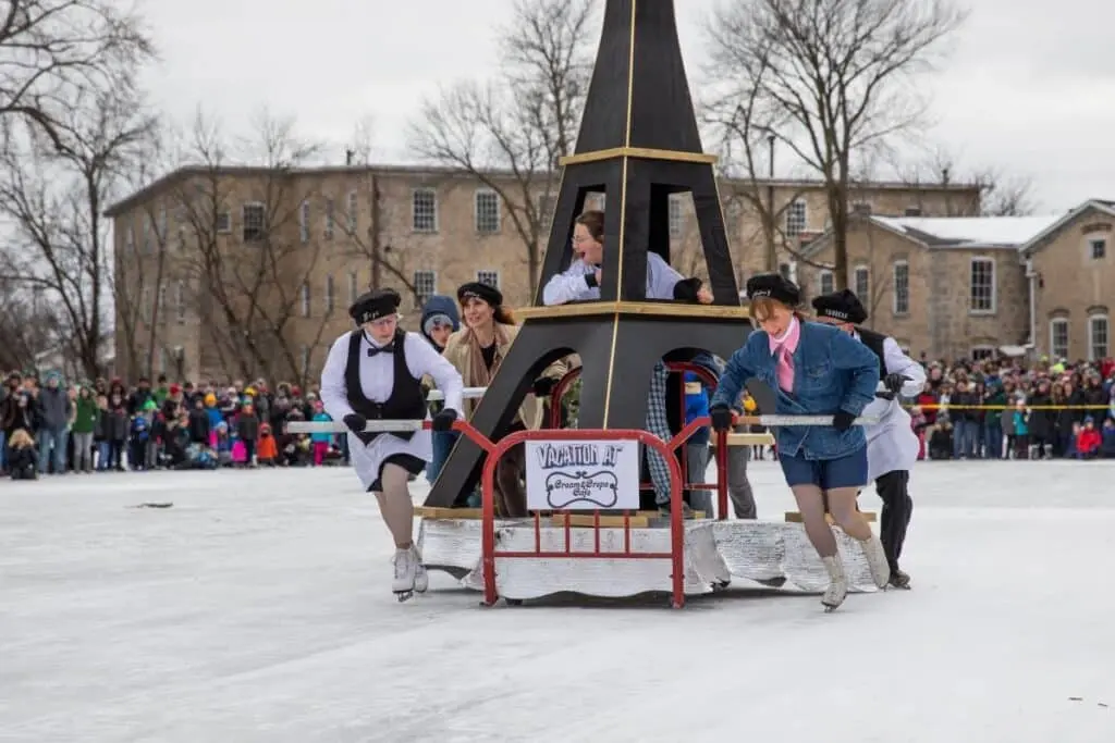 A group of women skaters pushing asled that looks like the Eiffel Tower while there are onlookers at A Winter Stroll Through Cedarburg event