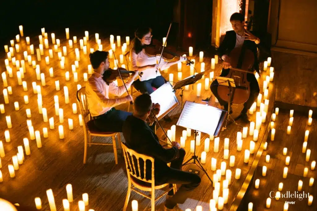 A group of four violinists sitting in half circle and playing some music with music book and surrounded by many lighted candles.