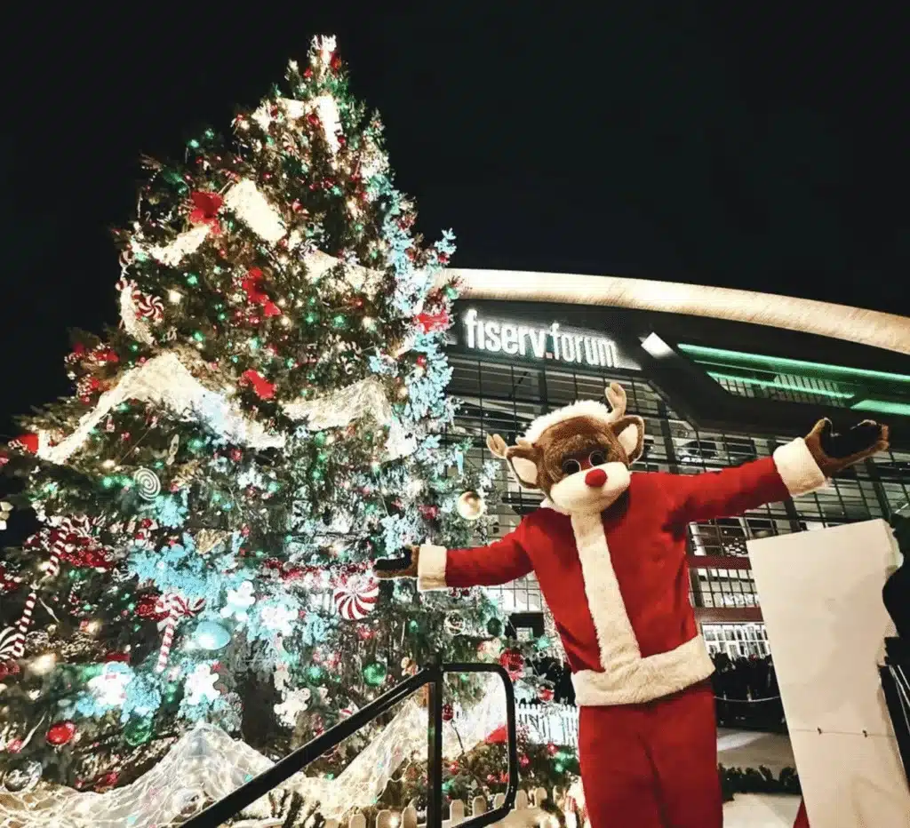 A Christmas deer in Santa Claus suit posing beside a giant Christmas tree in front of the Fiserv Forum.
