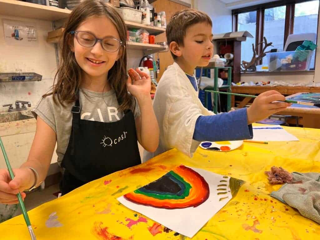 A girl painting a rainbow with a boy in blue shirt during art class