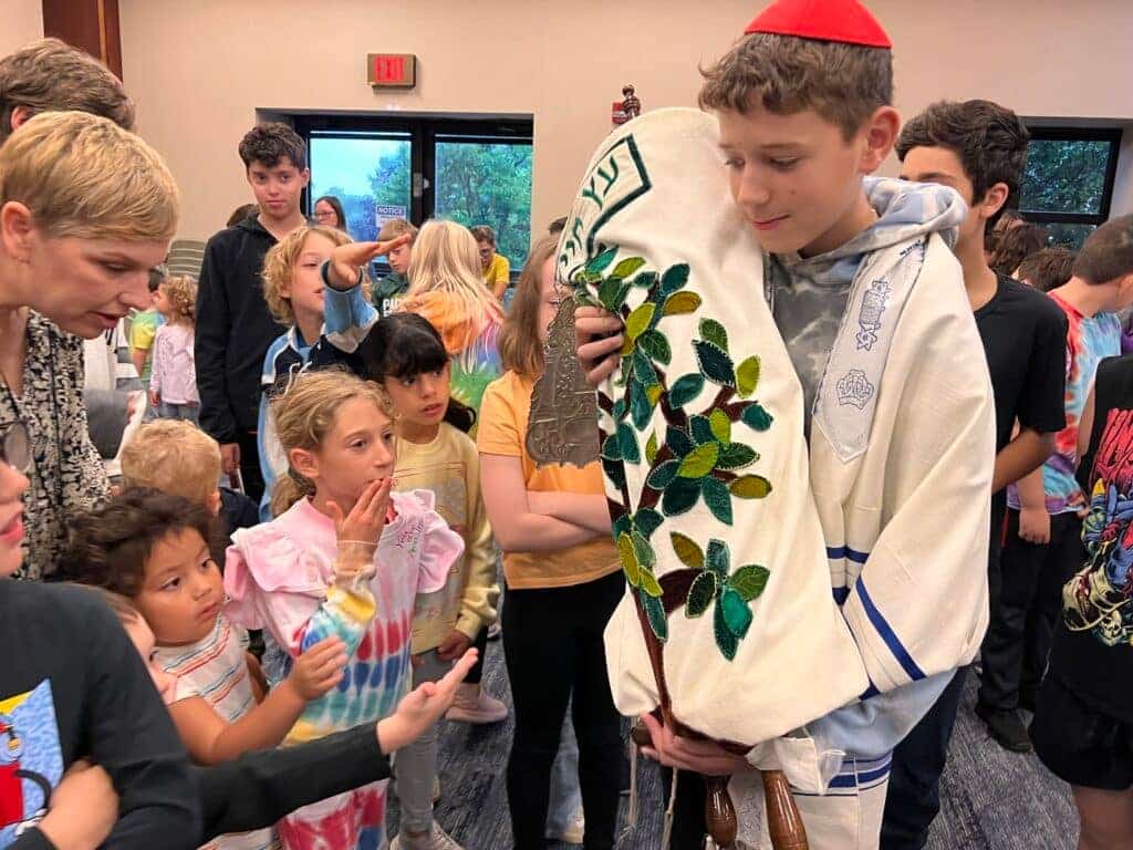 A boy wearing a Jewish parayer shawl and red kippah and holding a torah with decorative cover