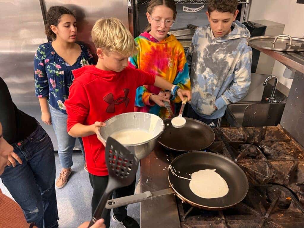 A teenage boy cooking pancakes and holding a bowl while some of his classmates are looking at him