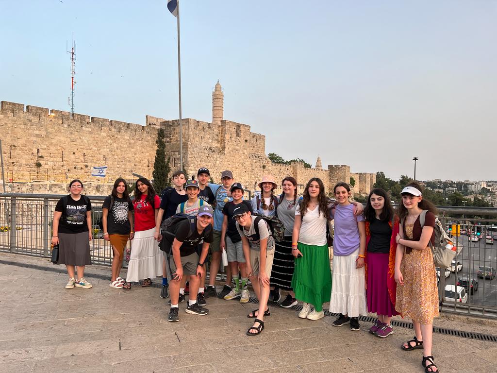 A group of teenages posing for a camera with an old city wall and the flag of Israel in the background