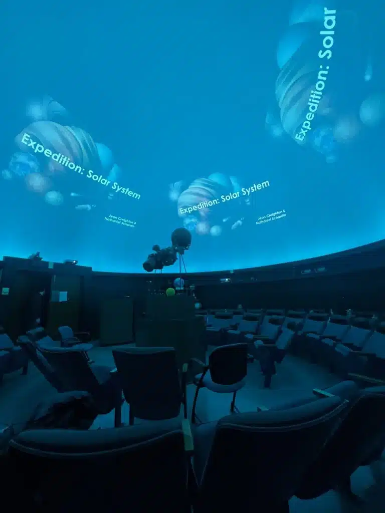 Blue-lit UWM Manfred Olson planetarium dome displaying “Expedition: Solar System” with seating and star projector below.