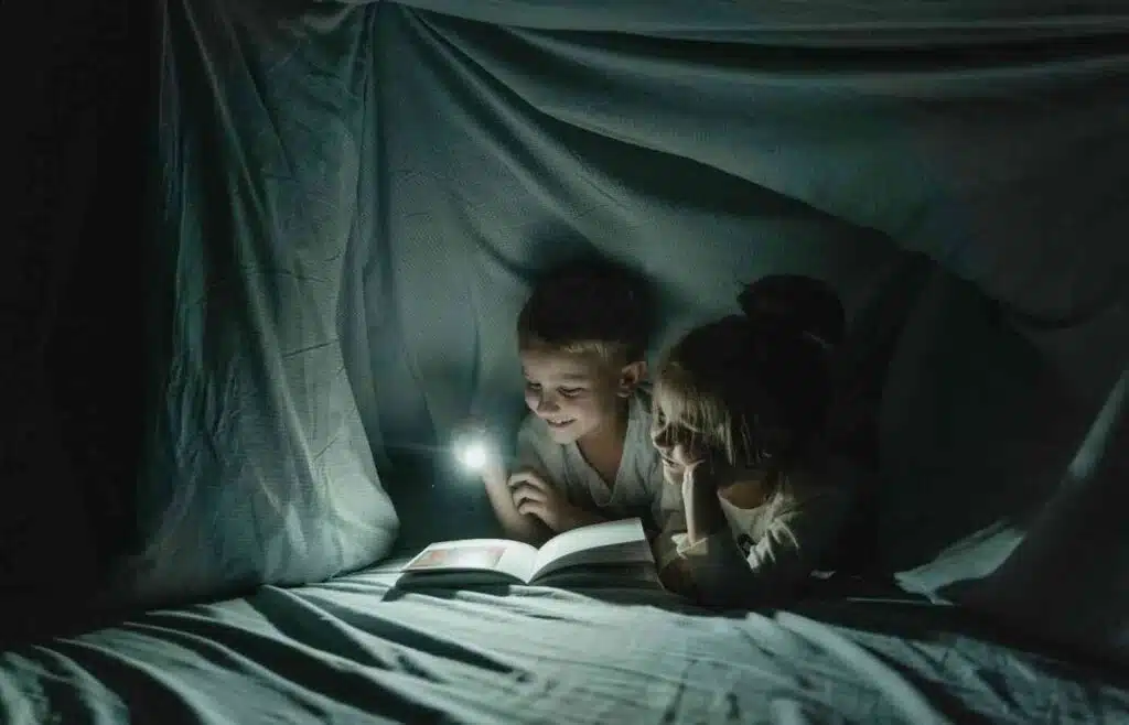 Two children reading with a flashlight inside a cozy homemade fort.