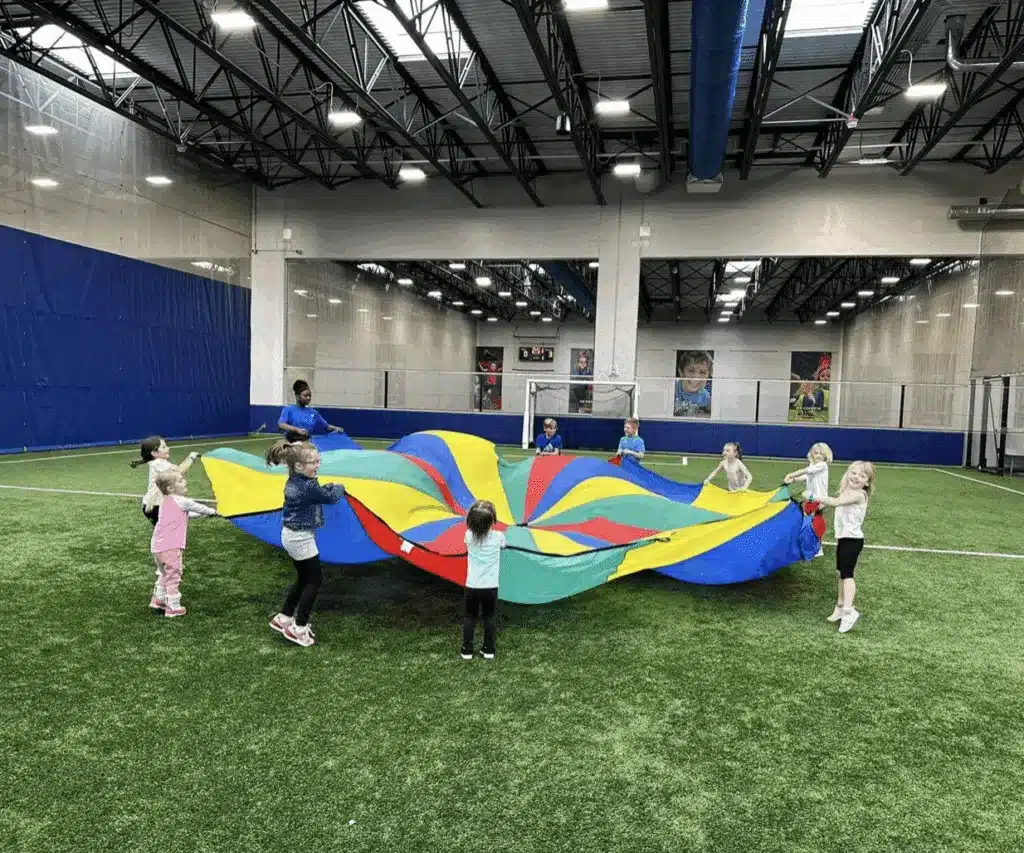 Ten kids are holding a multi-colored parachute in the middle of indoor Milwaukee Yard playground.