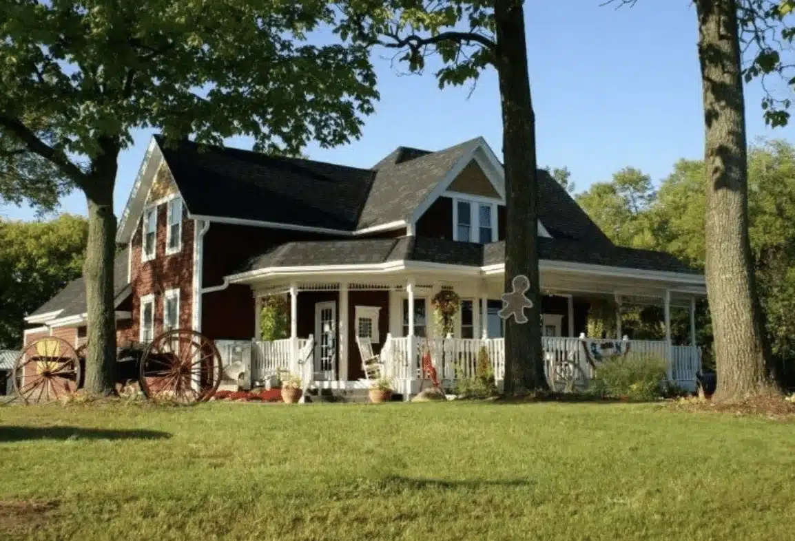 A quaint farmhouse with white porch  and brick red siding, white windows and surrounded by three trees.