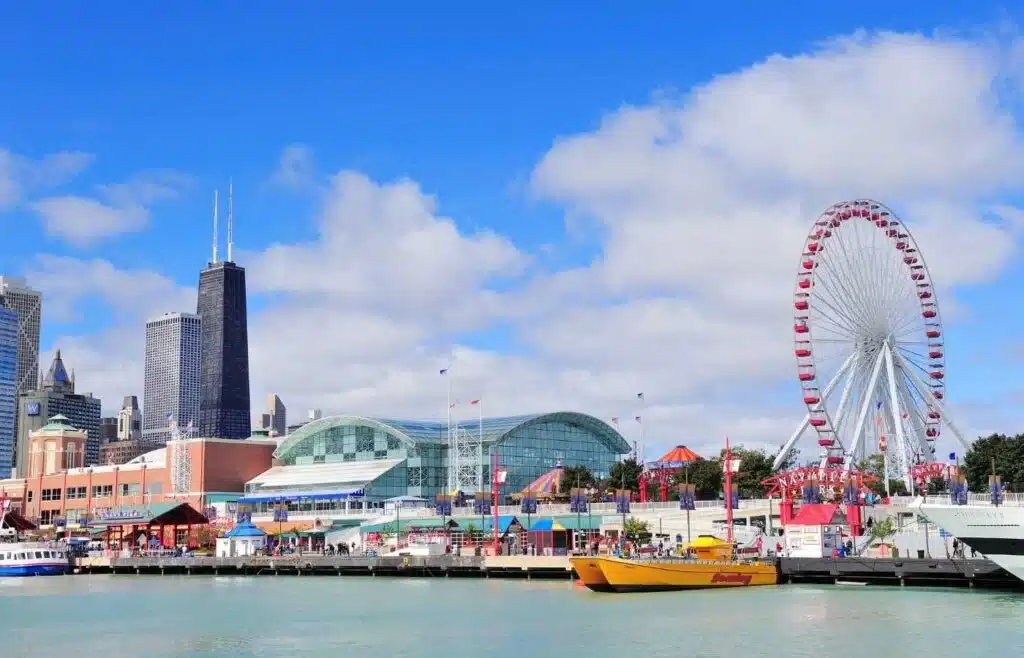 A waterfront view of Navy Pier in Chicago featuring the Centennial Wheel, lakefront attractions, and the city skyline under blue skies.