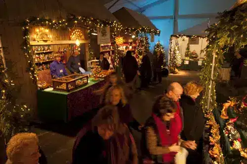 A street market selling holiday themed food, decor and gifts with stalls decorated with Christmas lights and pine tree branches