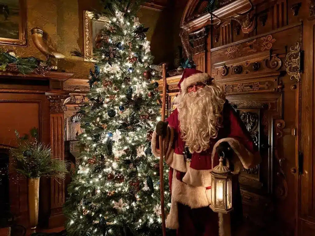 Father Christmas standing beside a Christmas tree and holding a lantern in his hand and cane on the other 