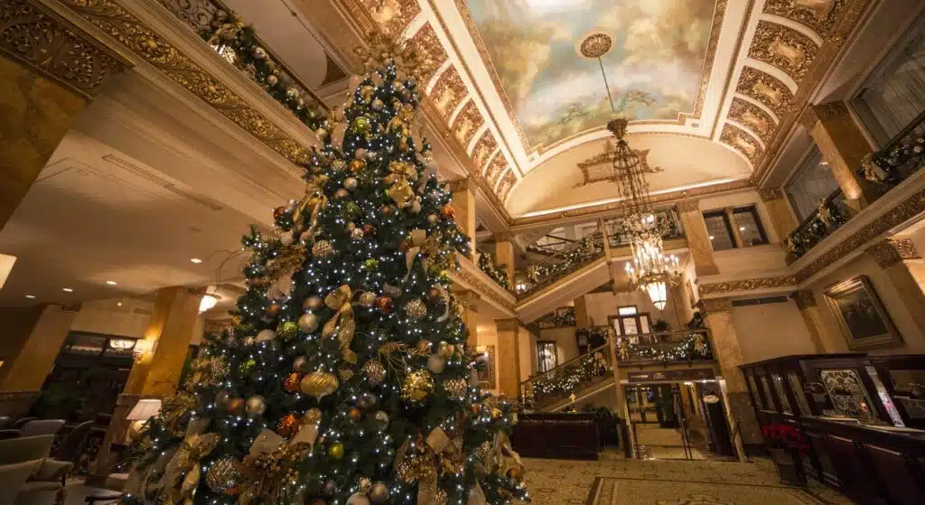 A well decorated Christmas tree with golden balls and ribbons inside a lavish hotel with painted ceilings and crystal chandelier.