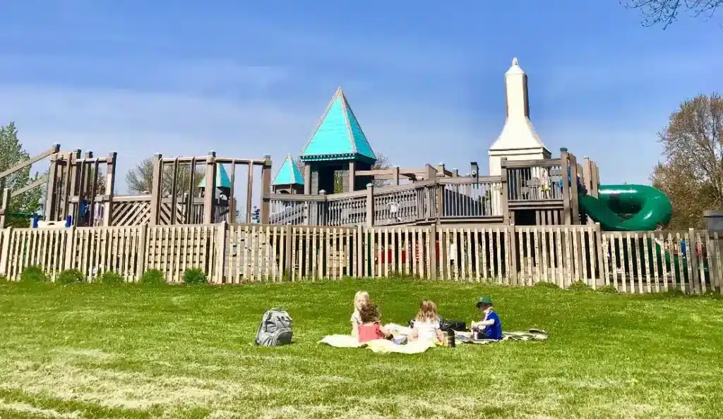 Children sit on a picnic blanket in the grassy field below Possibility Playground, with the large castle-themed play structure 
