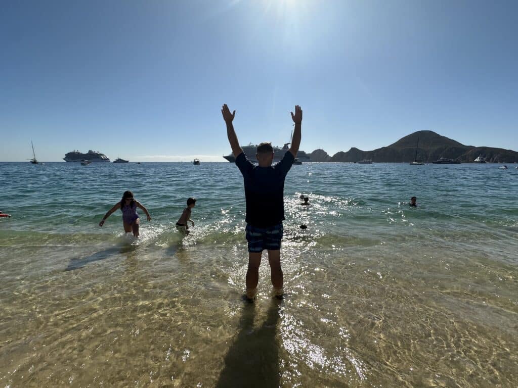 A vibrant beach scene at Pueblo Bonito Los Cabos with children playing in the calm waters, a man standing with arms raised, and scenic hills in the background.